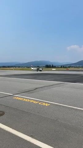 A small white airplane with red and yellow accents is on an asphalt runway under a clear blue sky with a few scattered clouds. The airplane is facing left and appears to be moving. In the foreground, the runway has yellow markings that read "NO PARKING." In the background, there are rolling green hills or mountains. The video is shot from a low angle, looking towards the airplane.