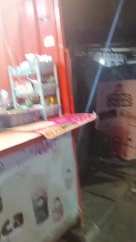 A counter is set up in front of a red corrugated metal wall. On the counter is a white pitcher with a brown liquid, a yellow fan with a dispenser of straws, and a blue bucket. Behind the counter, a menu board lists "FRUIT SERIES" and "MILK TEA SERIES". The front of the counter is decorated with cartoon images of a girl with pigtails and text that reads "teh oca".