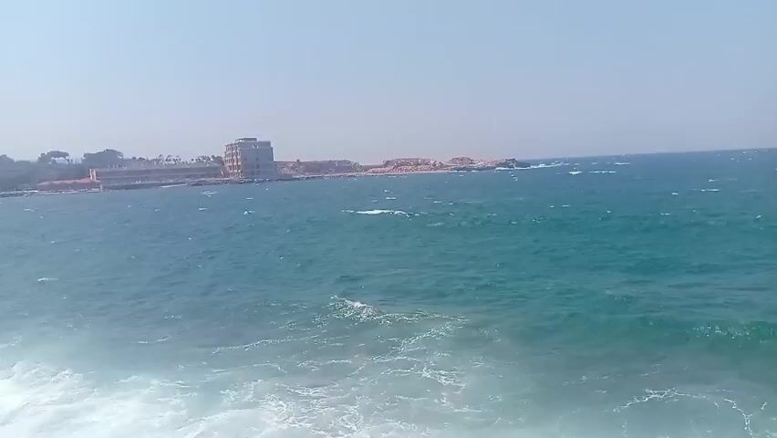 The video captures a view of the sea and a coastline with buildings. The water is a deep blue-green and appears somewhat choppy with waves crashing against the rocky shore. On the coastline, there are several buildings of varying sizes and architectural styles, some appearing to be hotels or residential structures. Trees and other vegetation are visible behind the buildings. The sky is clear and bright, suggesting a sunny day. The camera appears to be moving slowly, giving a panoramic view of the scene. The overall impression is that of a coastal town or resort area on a windy day.