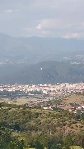 The video presents a static, panoramic view of a landscape encompassing a town, mountains, and hills, likely taken from a high vantage point overlooking a valley. In the middle ground, a densely built-up town or city is visible, characterized by numerous multi-story buildings. Beyond the town, a prominent, forested mountain rises. On the face of this mountain, a very large, bright white cross stands out against the green and grey hues of the hillside. The background features distant, hazy mountains under a sky with scattered clouds. In the foreground, lush green hills and sparse trees are visible, framing the view of the valley below. The scene is well-lit, suggesting daytime, possibly morning or afternoon. There is no visible text or human activity within the frame. The location context provided indicates this scene is in Selishte, Bulgaria.