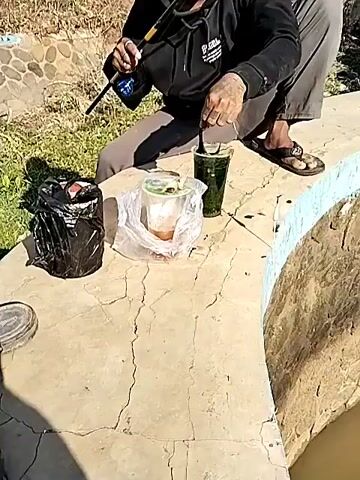 A person wearing a hooded black sweatshirt and grey pants is crouching down on a concrete ledge, holding a fishing rod. A black plastic bag with trash is to the left of the person. Next to that is a clear plastic container with food inside, and a green cup with a straw. The person appears to be fishing in a body of murky water that is contained by a stone wall. The wall has a blue and yellow trim along the top. Stairs made of concrete and painted blue and yellow are visible on the left side of the frame. The video pans across the water and up a steep, narrow channel with stone walls leading to a dark opening. A rope hangs down from above into the water.