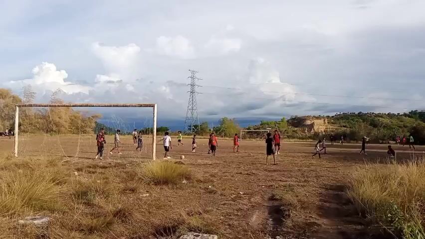 The video shows a group of people playing soccer on a dirt field. The field is covered in dry grass and is uneven. There are two soccer goals on opposite sides of the field. In the background, there are trees, hills, and a power line tower. The sky is cloudy. The people are wearing casual clothes and appear to be enjoying themselves. Some are kicking the ball, while others are running and chasing it. There are also some spectators sitting on the side of the field.