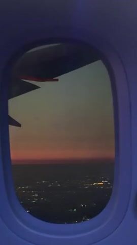 A view from an airplane window shows a city at night. The city lights twinkle below, while the sky is tinted with hues of orange and gray at the horizon. A portion of the airplane wing is visible at the top left of the frame. There is indistinct speech in the background.