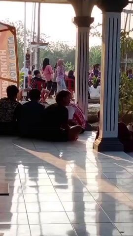 The video shows a gathering of people outdoors, possibly at an event or celebration. In the foreground, four individuals are seated on the tiled floor, facing away from the camera. Their backs are to the viewer, obscuring their faces. The flooring appears to be tiled and reflective, with sunlight casting distinct shadows. Behind them, a group of people are standing and interacting. Several individuals are visible, including women in colorful clothing and headscarves, and some men. A banner with Indonesian text is partially visible on the left side of the frame. The background suggests an outdoor setting with trees and a bright sky, indicating it is daytime. The overall atmosphere seems relaxed and social.