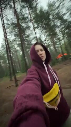 A young woman in a maroon hooded sweatshirt and a yellow fanny pack spins around in a forest. She is smiling and her eyes are closed. The ground is covered in dirt and pine needles. Tall trees with green foliage surround her. In the background, there are tents and orange lights, suggesting a campsite or festival. The lighting is dim, indicating it might be evening or overcast.