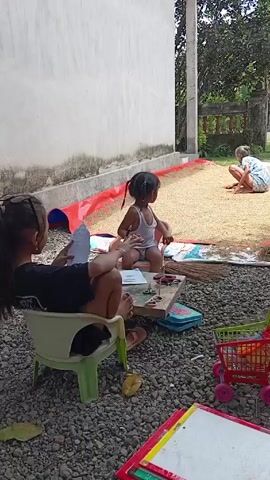 The video captures a scene of children and an adult engaged in different activities in an outdoor space. Two young girls are seated at a small table, which is covered with art supplies and a box. One girl sits in a green and white chair while the other is on a small stool. They appear to be drawing or crafting. A toy shopping cart sits near the table, filled with various items. In the background, an adult is squatting or sitting on a layer of what looks like grain spread out on a mat. The scene is situated near a white wall, with a gravel-covered ground, and lush greenery is visible in the background. The setting suggests a rural or semi-rural environment with everyday activities taking place.