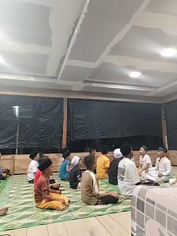 A group of young boys are sitting on a green and white mat on the floor. They are all facing the same direction, some are looking at a boy in a white garment who appears to be teaching or leading them. The room has wooden walls and a drop ceiling with some unfinished sections. Black sheeting is hung on the walls behind the boys. The lighting appears to be from overhead fixtures.