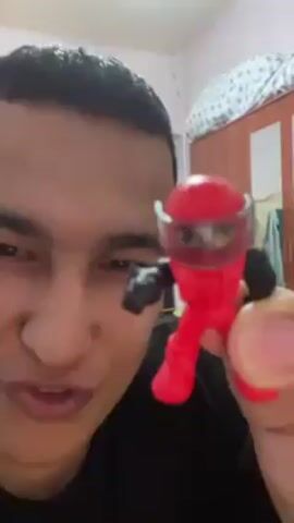 A close-up shot shows a man holding a red toy figure of a person in his right hand. The toy figure has a helmet with a transparent visor and black gloves. The man looks at the camera and moves the toy up and down in front of his face. He smiles at the end of the video. The background is blurry but appears to be a room.