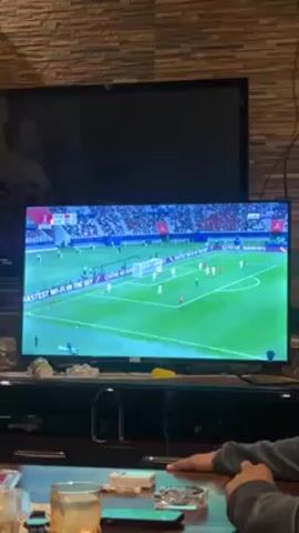 The video shows a person sitting at a table watching a soccer match on television. The television is showing the game with the score Morocco 0 - Croatia 0. The person is sitting in front of the television and is looking at it. There are other objects on the table, including a glass of water and a pack of cigarettes. The room is decorated with a stone wall. The referee of the soccer game is on the television screen, holding his arm out to the side.