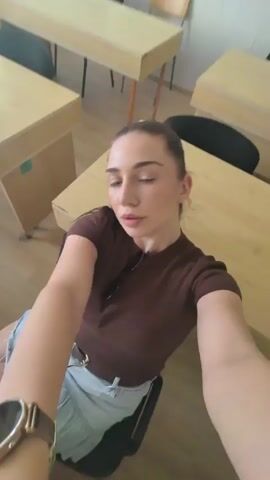 The video begins with a close-up shot of a young woman with dark hair pulled back, wearing a brown short-sleeved shirt and light denim shorts. She is seated in a chair at a wooden desk in what appears to be a classroom or office environment, with other desks visible in the background. The camera angle is from a high perspective, looking down at the woman. She looks directly into the camera and smiles. The scene then shifts to a slightly different angle, still looking down at the woman. Her hair is partially obscuring her face. The video continues with varying close-up shots of her face and upper body, with the camera angle constantly shifting and moving, sometimes appearing to be handheld and shaky. Sunglasses are visible hanging from her shirt. The background remains the same, showing wooden desks and chairs. The woman occasionally moves her head and shifts her body, and her facial expressions vary from a smile to a more neutral gaze.