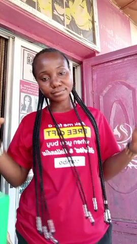 A young woman with dark skin and braided hair stands in front of a beauty salon, wearing a red t-shirt with white text, black pants, and a wrist bracelet on each wrist. She raises and lowers her hands above her head, then makes a gesture with both hands as if rejecting something, then smiles slightly while looking at the camera. The salon has a glass sliding door and a pink door next to it, both with signs. A small green container is partially visible to the left of the woman.