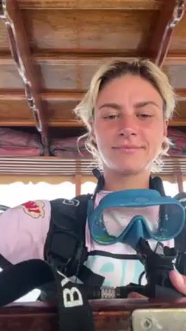 A woman with short blonde hair is sitting down wearing a scuba diving vest. She has a scuba mask around her neck. The woman has a black scuba diving regulator in her hands. She puts the regulator in her mouth, takes a deep breath, and then takes it out of her mouth. The video appears to be filmed on a boat, as wooden roofing can be seen above the woman. The boat motor is heard in the background.
