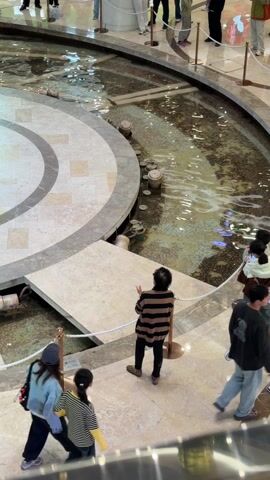 The scene is a high-angle view of the interior of a large, modern shopping mall with a large, circular decorative fountain. A group of people stand near the fountain, looking at the water. The fountain is inlaid with patterned tilework and appears to contain coins in the water.  A small marble platform extends over the water, and the area is partially cordoned off with ropes.  The people present include a woman in a denim jacket and baseball cap, a child in a striped shirt, and a person in a black and brown striped top. Other mall patrons can be seen walking nearby.  The floor is tiled. The overall lighting is bright and well-lit.