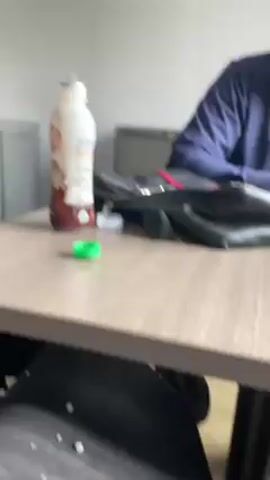 A slightly out-of-focus indoor shot shows a man sitting at a table. He wears a dark blue jacket and appears to be looking to his right. On the table is a chocolate milk bottle, a green bottle cap, and a black bag with red accents. The table has a light wood grain finish, and the walls are painted a neutral grey.