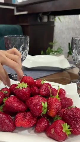 The video shows a hand picking up a strawberry from a white plate laden with fresh strawberries. The scene is set on a table with dining arrangement, including crystal glasses, silverware, and white plates. The background suggests a restaurant setting with dark wood accents and a hint of greenery. The focus is on the vibrant red strawberries and the dining setting.