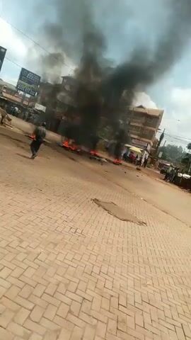 The video shows a street scene that appears to be a protest or riot. There are fires burning in the street, with thick black smoke rising into the air. A group of people are standing near the fires, and some are walking around the area. The street is paved with brown bricks, and there are buildings on either side of the street. There are also billboards and utility poles visible. A motorcycle with two people on it navigates around the burning areas. The overall environment suggests a scene of civil unrest or demonstration.