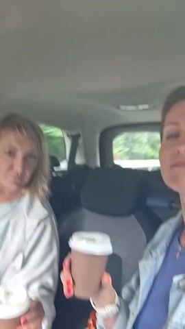Two women are sitting in the back seat of a car, holding disposable coffee cups. The woman on the left has blonde hair and is wearing a light blue jacket. The woman on the right has short brown hair and is wearing a blue jacket over a purple shirt. Both women are smiling and looking at the camera. The car appears to be in motion, with trees visible outside the windows. There are some containers of food on the seat in front of them.