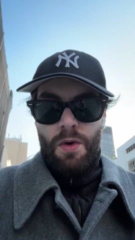 A person wearing sunglasses, a grey coat, a black turtleneck sweater, and a New York Yankees baseball cap is looking directly at the camera outdoors. The person's mouth is open slightly. Buildings are visible in the background. The sky is blue and clear.