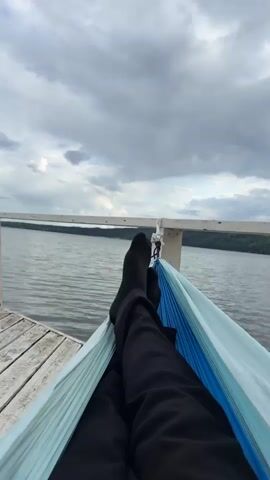 The video shows a first-person view of someone lying in a hammock. Their legs, clad in black pants and black socks, are visible in the foreground. The hammock is a light blue and teal color. The hammock appears to be suspended from a white railing that runs along the edge of a wooden dock. Beyond the dock and railing, a body of water stretches out, with a distant shoreline visible under a cloudy sky. The water is calm, with gentle ripples. The clouds are a mix of gray and white, suggesting an overcast or potentially stormy day. The perspective is looking upwards towards the sky and out over the water.