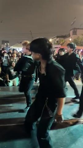A group of young people in black clothing are dancing on a stage outdoors at night. The person in the foreground is a male with dark, curly hair, wearing a black shirt, black pants with a chain, and black shoes. He is in motion, with his arms outstretched. Behind him, other dancers are also performing. A crowd of spectators is visible behind the stage, some sitting and some standing. Tents and buildings are in the background, illuminated by lights. The stage is lit with purple and blue lights, casting shadows.
