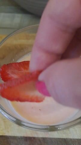 A person is adding sliced strawberries and basil leaves to a clear glass bowl filled with a light-colored, creamy substance. The bowl is on a wooden surface. The person's hand, which has pink nail polish, is placing the strawberries and basil carefully. The view is a close-up, focusing on the bowl and the action of adding the ingredients.