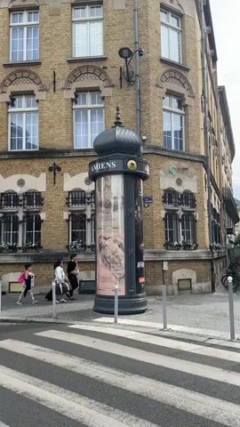 The video shows a street scene with a prominent cylindrical advertising pillar in the foreground. The pillar is black and adorned with posters and a decorative dome-like top. The building behind it is made of yellow brick with numerous windows. Several people are visible walking past the pillar and on the sidewalk. In the background, there are more buildings, trees, street signs, and a crosswalk. One sign indicates a pedestrian zone. A man walks towards the camera in the foreground. The overall environment suggests an urban setting on a cloudy day.