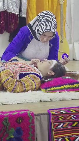 In the video, a woman wearing a blue jacket and a black and white headscarf is applying something to the face of a young person who is lying down. The young person is wearing a colorful, traditional-looking outfit with intricate embroidery and is lying on a white rug. A pink and yellow pillow is under the young person's head, and brightly colored woven mats are visible in the foreground. In the background, there are draped fabrics in yellow and silver, and a stringed instrument that appears to be a lute or a balalaika. The woman is holding a small purple container in her right hand and a brush or applicator in her left hand, which she is using on the young person's face. The young person's eyes are closed, and their face is turned upwards. The overall scene suggests a cultural or artistic performance or preparation.