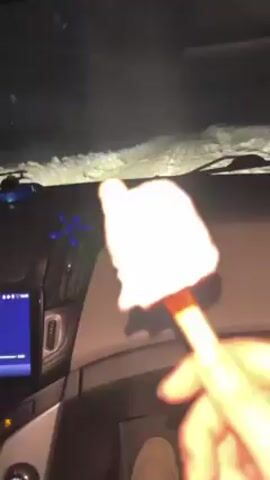 A person in a car holds up a partially eaten ice cream bar on a stick. The ice cream is melting. A blue digital display on the car dashboard is illuminated. There are decorations on the dashboard, including a beaded hanging ornament. Outside the car, snow is piled high on the windshield. Music is playing.