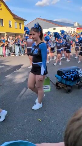 The video shows a group of cheerleaders in blue and black outfits participating in a parade. The cheerleaders are of various ages, with some younger girls and older teenagers. They are carrying pom-poms, some of which are blue and silver. One of the cheerleaders in the foreground is pulling a small, grey wagon filled with more blue and silver pom-poms. The cheerleaders are walking down a street lined with buildings and spectators. The sky is blue with some clouds, and the sun appears to be shining. There is a yellow building with a sign that reads "Buttstädter Volks Bäckerei" in the background. The overall atmosphere appears festive and celebratory.