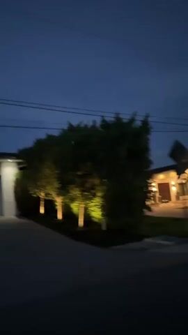 The video shows a view from a moving vehicle at dusk. The camera pans across a suburban street lined with houses and palm trees. The houses are illuminated with exterior lights, casting a warm glow on the white facades. A white SUV is parked in the driveway of one of the houses. The sky is a deep blue, with a few wispy clouds. The overall impression is one of peaceful tranquility and suburban affluence.