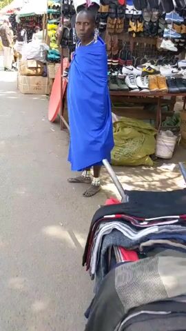 Here's a concise description of the video: 

The video shows an outdoor street market scene in what appears to be an African country. Several vendors are set up along the sidewalk, displaying goods for sale. One vendor, a woman in a bright blue wrap, stands near a display of shoes and sandals. Another vendor's display is backed by a red AzamTV satellite dish. People are walking by, seemingly browsing or shopping. The environment appears to be bustling with activity and commerce.