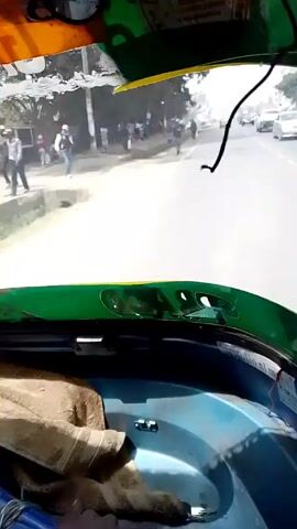 The view from the interior of a green and yellow auto rickshaw, also known as a tuk-tuk, shows a bright, sunlit road ahead with cars and pedestrians. On the left side of the road, a group of people stand near some trees, some are walking along the side of the road. Other cars are visible driving in the distance.  Inside the tuk-tuk, a blue passenger area is seen with a brown cloth on the seat. The tuk-tuk is in motion, moving forward on the road.