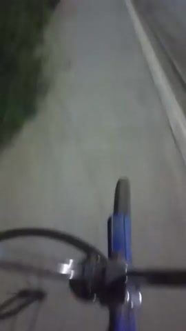 The video shows a first-person perspective of someone riding a bicycle on a paved sidewalk next to a street. The lighting suggests it is either dusk or nighttime, with streetlights or other artificial illumination visible. Green foliage is present on the left side of the sidewalk. The road on the right appears to be empty. A traffic light is visible in the distance, showing a red signal. The camera is mounted low, focusing on the front wheel of the bicycle and the path ahead.
