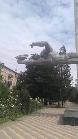 The video shows a statue of a swimmer with arms extended, seemingly mid-stroke. The statue is mounted on two tall, concrete pillars. The pillars are part of a monument that appears to be in a park or public area. A paved walkway runs in front of the monument. Trees and bushes line the walkway. A building is visible in the background to the left. A person is walking on the walkway. The sky is cloudy. The video pans slightly to the right, showing more of the monument and the surrounding environment.