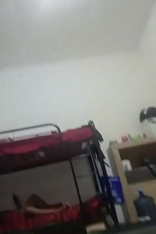 This is a video captured on a phone, likely indoors, with a somewhat blurry and shaky quality. The scene appears to be a shared living space, possibly a dormitory or a budget accommodation, given the presence of bunk beds.

In the foreground, a person's legs, clad in black pants, are visible, suggesting they are seated or lying down, facing away from the camera. A laptop or tablet is positioned in front of them, displaying an image of a potted plant with white flowers and a person wearing a cap and an orange shirt.

The background reveals a bunk bed with red bedding, and a person is lying on the lower bunk, partially visible. Further back, a tall, light-colored cabinet with shelves holds various items, including a blue plastic container, a large water dispenser jug, and other unidentifiable objects. A dark-colored shelf unit is also present, topped with what looks like a white box and a black hat.

To the right, a wooden stool with a red object on it is visible. A green cabinet is seen to the left of the frame. The floor is tiled in a light color. The overall lighting suggests it could be daytime, but the indoor setting makes it difficult to ascertain the time of day definitively. The atmosphere seems relaxed, possibly indicating a casual downtime activity like watching something on a device. No specific events or emotions are strongly conveyed, beyond a sense of everyday life. There is no visible text in the video. The location context of Surabaya, East Java, aligns with a tropical climate, but weather conditions are not discernible from the footage.