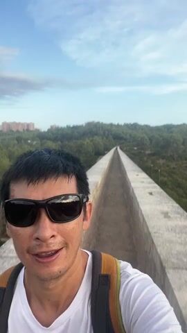 A man with short black hair and sunglasses smiles at the camera while standing on a concrete aqueduct. He is wearing a white t-shirt and a backpack with orange and yellow straps. The aqueduct extends into the distance, lined with trees and hills. The sky is blue with scattered clouds. The man appears to be on a hike or excursion.
