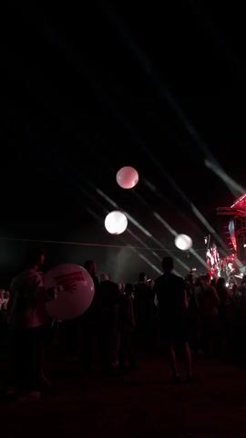 The video is a vertical shot of a night event, possibly a concert or festival. The background is dark, with bright spotlights cutting through the air from right to left, creating visible beams. In the distance, a stage with lighting structures is visible on the right. In the foreground, a crowd of people are silhouetted against the lights, facing the stage. A large, white balloon with red lettering, possibly a logo, is held by a person on the far left. Three glowing, spherical objects, likely illuminated balloons, are floating in the sky, two pinkish-white and one white. The overall atmosphere appears to be energetic and festive.