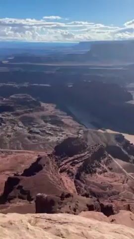 The video displays a wide-angle, sunlit, daytime view of a vast canyon landscape. A river runs through the canyon floor, its color varying from muddy brown to reflective silver due to the sunlight. The canyon walls are a mix of red and brown rock formations, with layers of plateaus stretching into the distance. The sky is blue with scattered white clouds. A dirt road is visible winding along a portion of the canyon floor. The camera appears to be positioned at a high vantage point. The wind is audible in the background.