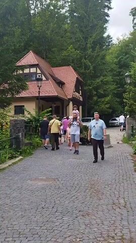 A group of people are walking on a cobblestone path in front of a building nestled amongst trees. A man in a blue t-shirt and dark pants walks towards the camera. To his left, a man with a child on his shoulders walks away from the camera, followed by a woman in a red dress. Other individuals are visible in the background, and parked cars are present. The building appears to be a house with a red tile roof and timber accents. The environment is lush and green, indicating a natural setting.