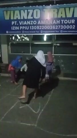 A group of people, including several children wearing Islamic attire, are gathered outdoors at night. A hopscotch grid is drawn on the ground in chalk. One person is actively playing hopscotch, while others observe or participate in the activity. In the background, there are buildings with signs, including one that reads "VIANZO TRAVEL." The lighting is dim, suggesting it is nighttime.