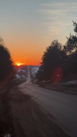 A static shot captures a sunrise over a winding asphalt road flanked by silhouetted trees. The sun is a vibrant orange orb touching the horizon, illuminating a distant town covered in snow. A dark-colored car drives away from the camera on the right side of the road. A second, smaller car is further up the road. The sky transitions from a pale blue at the top to a warm orange near the horizon. Bare patches of ground are visible at the roadside, indicating a partial thaw.