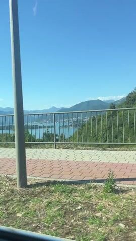 A stationary, medium-shot video captures a scenic view of a bay or large body of water with mountains in the distance. In the foreground, a metal fence runs horizontally across the frame, separating a paved walkway from the natural landscape. The walkway is patterned with reddish-brown and light-colored square tiles. Beyond the fence, the blue water is visible, dotted with boats, and the distant mountains are shrouded in a light haze. The sky is a clear, bright blue. The bottom of the frame shows the front of a dark-colored car, with its license plate partially visible. The overall impression is a bright, sunny day in a coastal or mountainous region.