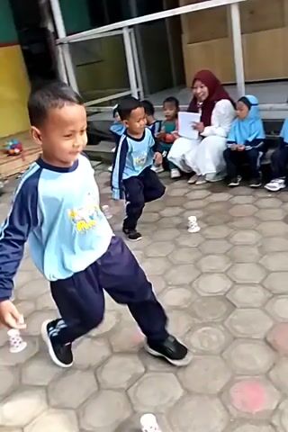 In this video, several young children are participating in an outdoor activity. The ground is paved with hexagonal tiles, and there are buildings visible in the background, suggesting a school or community setting.

The children are wearing school uniforms, consisting of light blue shirts with navy blue sleeves and pants. Some of the children are actively engaged in a game that involves running and interacting with small objects placed on the ground, which appear to be paper cups.

At the beginning of the video, a young boy in the foreground is looking to his right, seemingly focused on the activity. In the background, other children are also running and playing. As the video progresses, the perspective shifts, showing more of the playing field and the children's movements.

One child is seen running towards a set of cups, while another is in the process of picking up a cup. There are adults observing the children from the side, some seated and others standing. The overall atmosphere appears to be one of playful engagement and organized activity for the children. The lighting suggests it is daytime, with sunlight casting shadows on the ground.
