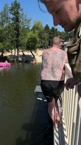 A man with extensive tattoos on his back and arms stands on a metal platform extending over a body of water. He is facing away from the camera and appears to be preparing to jump. He is wearing black swim shorts. To his right, a white railing runs along the edge of the platform. The water is dark and appears to be calm. In the background, there is a sandy beach with people visible, trees, and a bright blue sky. A pink inflatable object, possibly a toy, floats in the water to the left of the platform. Another person is partially visible in the upper right corner of the frame, looking down towards the man. The scene is set outdoors on a sunny day.