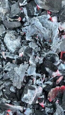 The video is an extreme close-up, top-down view of glowing charcoal embers and ash. The charcoal pieces are black and grey, with some showing bright red and orange glowing interiors, indicating they are hot. The ash is mostly a light grey color and appears dry and papery, covering parts of the charcoal. The texture of the scene is rough and uneven, composed of many irregularly shaped fragments. There are no people, animals, or other objects visible. The environment is solely the pile of burning charcoal.