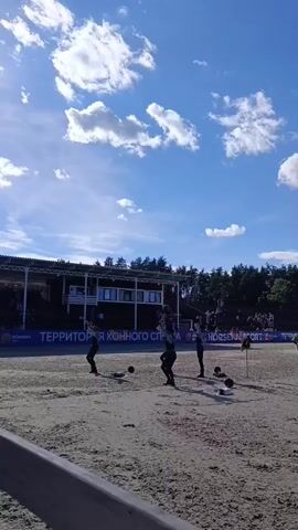 The video shows a sandy outdoor arena with three people standing in the center. The sky is blue with scattered white clouds. In the background, there is a covered grandstand with spectators and trees beyond. The three individuals are dressed in dark clothing and appear to be participating in some sort of performance or training. There are black balls on the ground near them, and one person is holding a ball. They are all facing the same direction, and their stances suggest they are engaged in a coordinated activity.