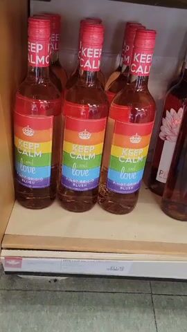 The video shows a shelf of wine bottles with rainbow-colored labels. The labels read "KEEP CALM and love" and "PINOT GRIGIO BLUSH". The bottles are arranged in rows on a shelf in what appears to be a store. The lighting is bright, and the background is blurred, suggesting the focus is on the wine bottles.