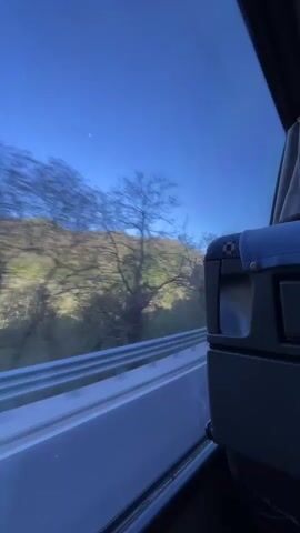 The video shows a view from inside a moving vehicle, likely a bus, through the window. Outside, a landscape of hills and trees blurs by. The sky is clear and blue, with a distant, pale object visible which could be the moon. Inside the bus, the seat and window frame are visible, providing a stationary point of reference to the moving scenery outside.