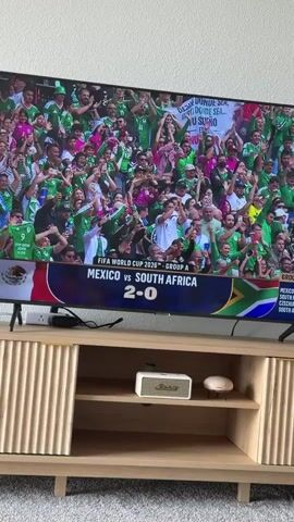 A wide-angle, eye-level shot shows a large flat-screen television displaying a soccer match. The screen shows a crowded stadium filled with spectators wearing green, and a scoreboard indicating "FIFA WORLD CUP 2026 - GROUP A MEXICO VS SOUTH AFRICA 2-0". The television is placed on a wooden media console with two open shelves. On the top shelf, there is a small white speaker and a small white object resembling a mushroom. The background is a plain wall.