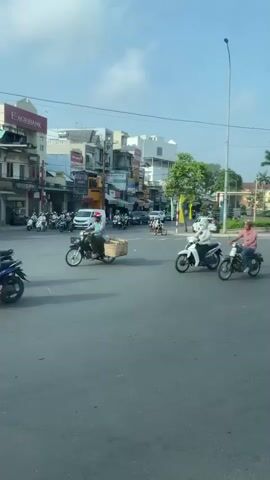 The video shows a busy intersection in Vietnam. There are many motorbikes and a few cars. The buildings are a mix of old and new, and there are many signs in Vietnamese. A green VinFast electric taxi passes in front of the camera. The intersection has a roundabout in the middle with Vietnamese flags, palm trees, and decorative plants. People on motorbikes can be seen crossing the crosswalk. The sky is blue with some clouds.